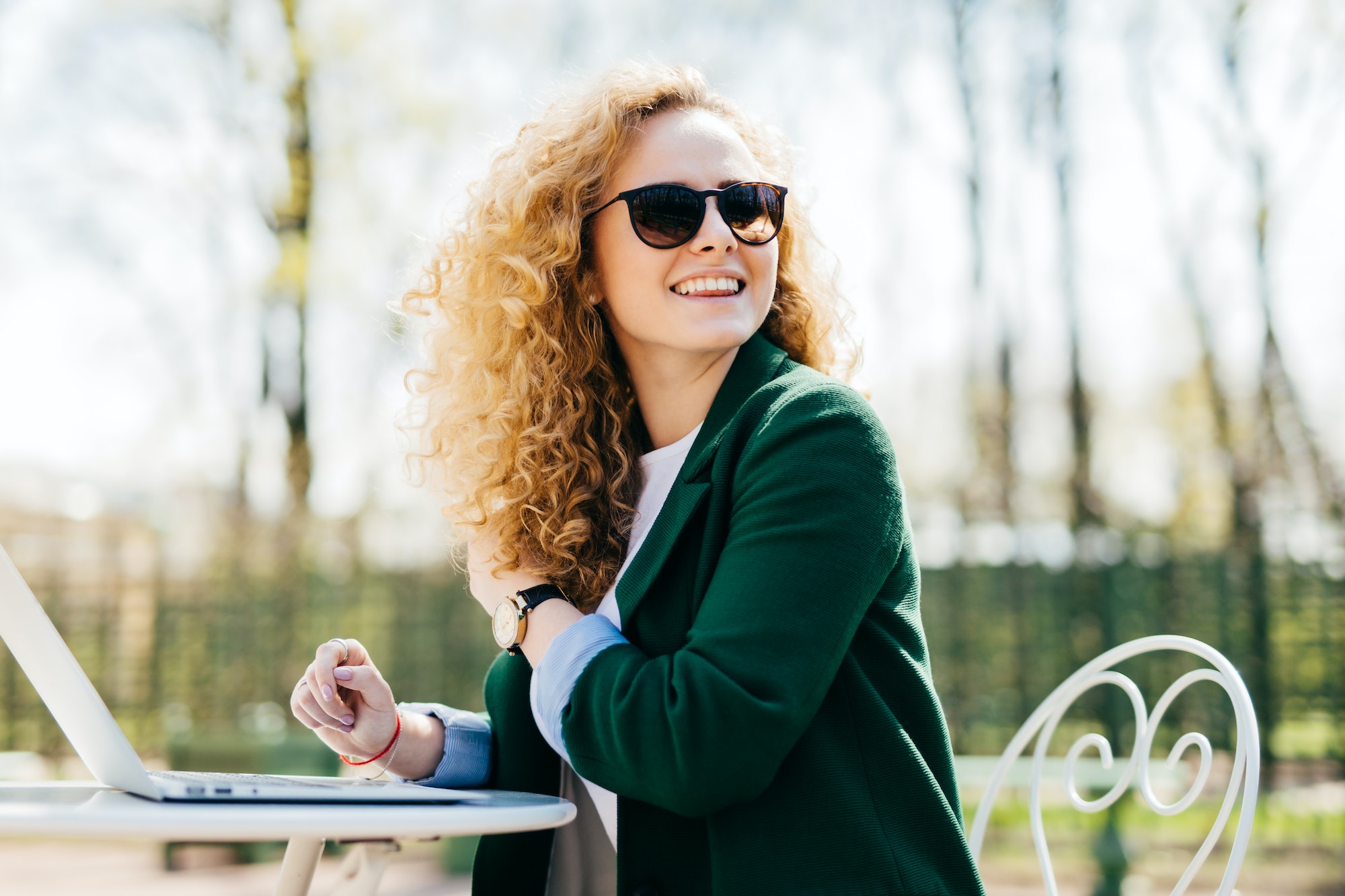 Happy stylish woman with curly light hair wearing sunglasses working with laptop outside in park