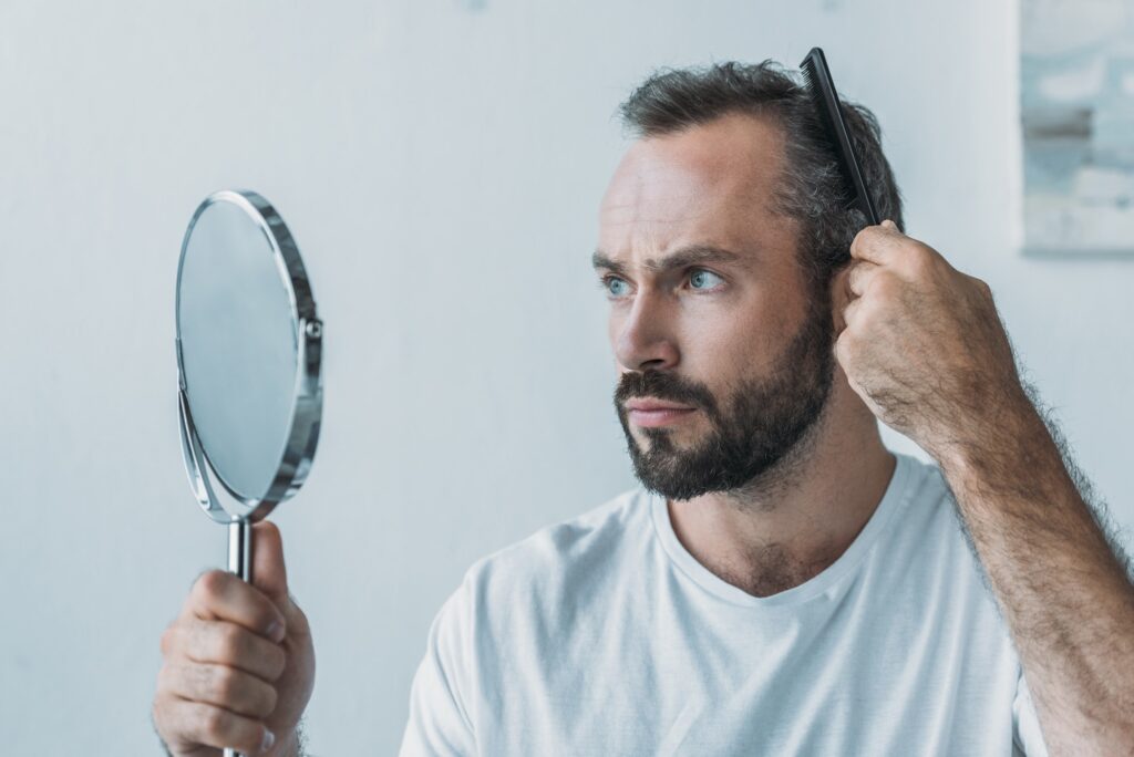 bearded middle aged man combing hair and looking at mirror, hair loss concept