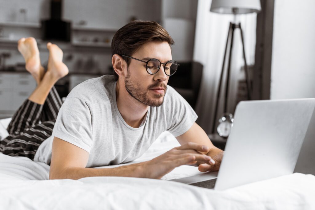 handsome young man in eyeglasses and pajamas using laptop on bed