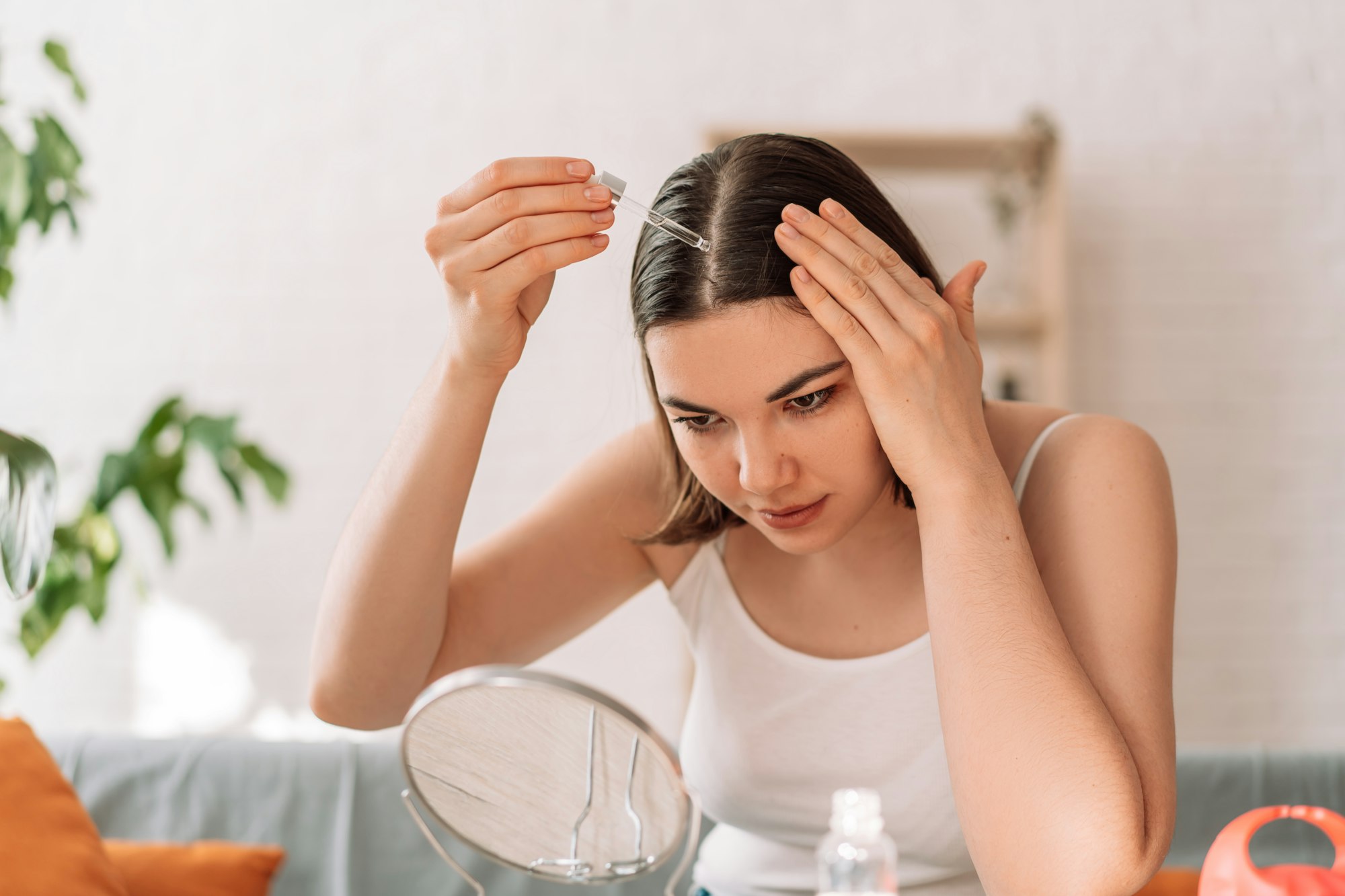 Lady sitting on the couch looks in the mirror and drips hair oil with a pipette on her hair after hair transplant