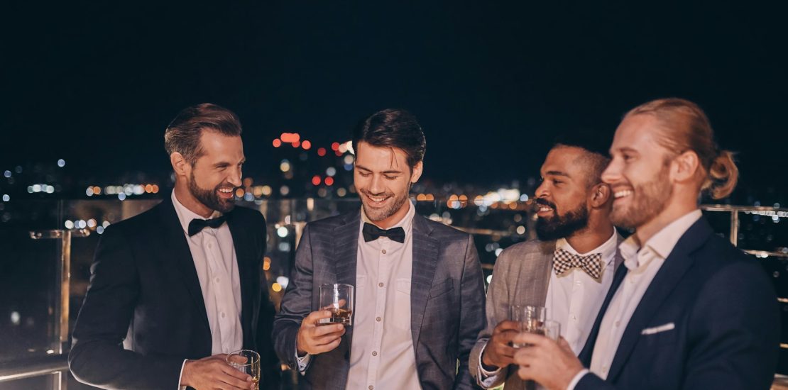 Group of handsome young men in suits and bowties drinking whiskey and smiling