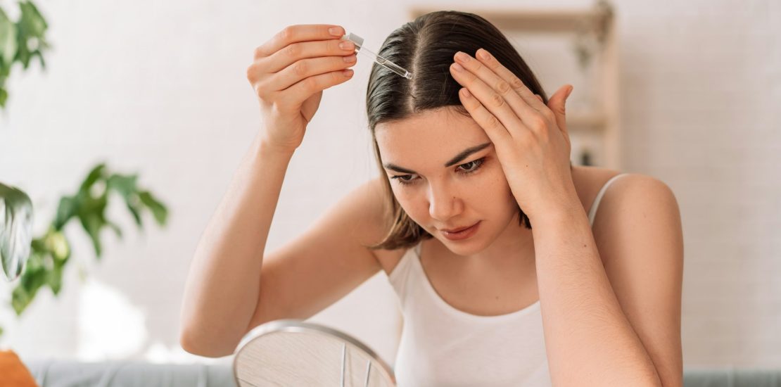 Lady sitting on the couch looks in the mirror and drips hair oil with a pipette on her hair after hair transplant