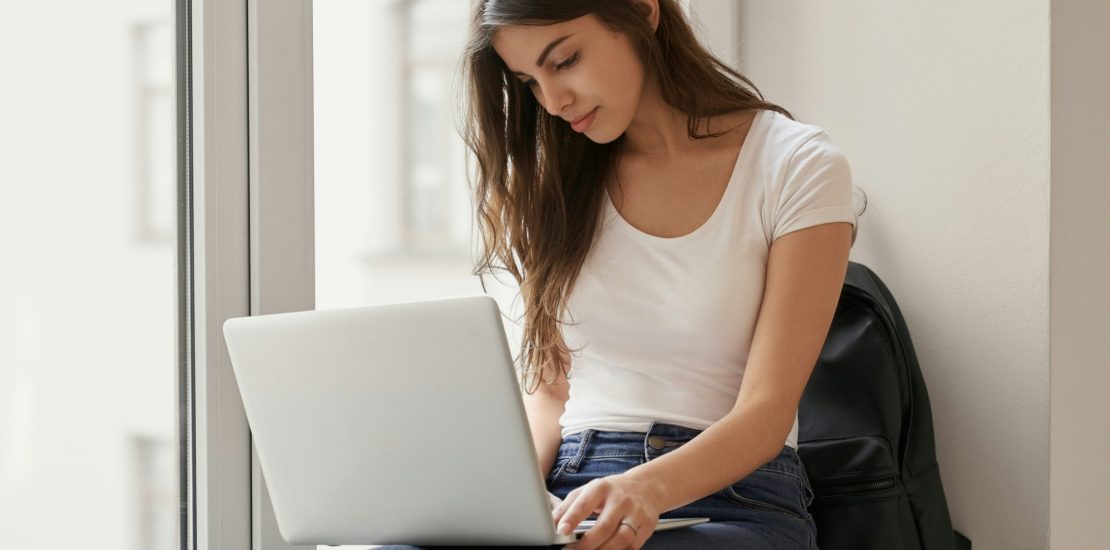 Pretty girl sitting on windowsill with laptop in university looking for advices for hyaluronic acid injections